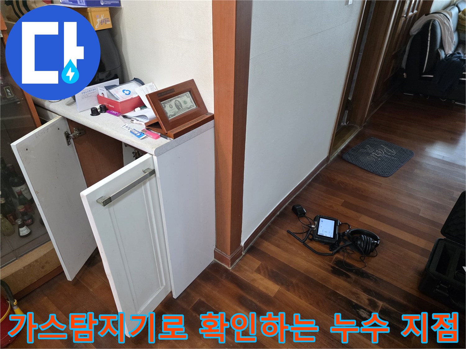 거실 바닥 근처에서 가스 탐지기를 사용하여 배관의 미세한 물샘 위치를 추적하는 모습입니다.
