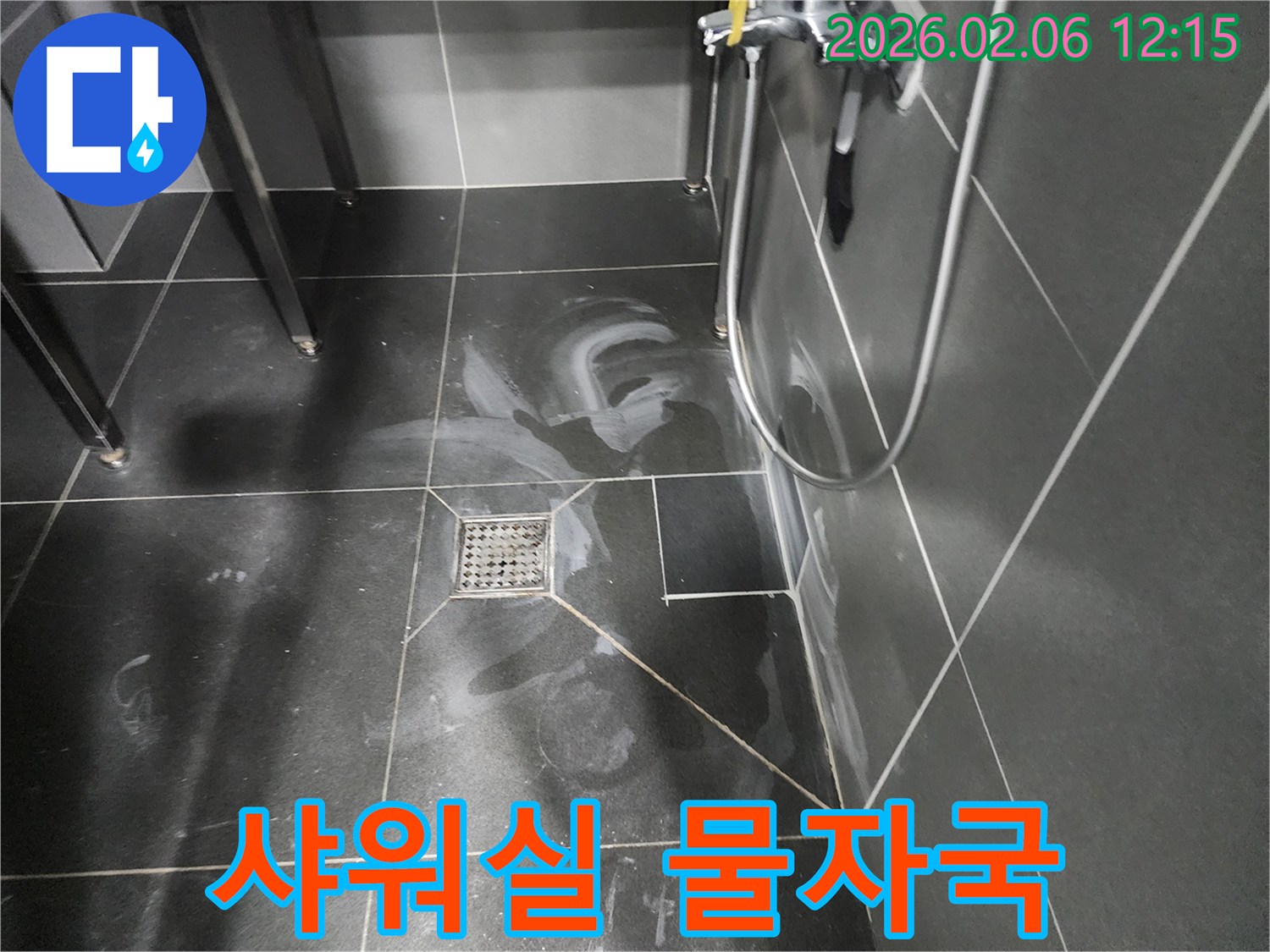 샤워 공간의 어두운 타일 바닥에 배수구와 함께 하얀 침전물과 물기가 얼룩져 있는 모습입니다.
