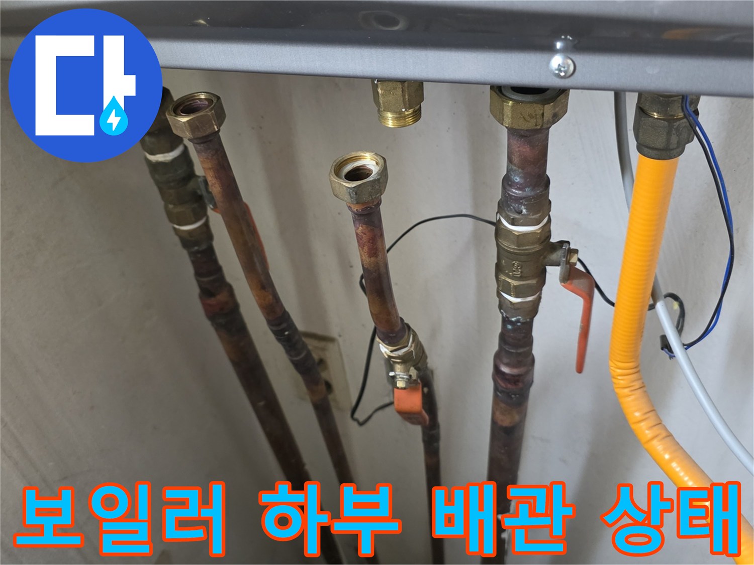 보일러 아래에 연결된 여러 개의 금속 관과 노란색 호스의 모습입니다.