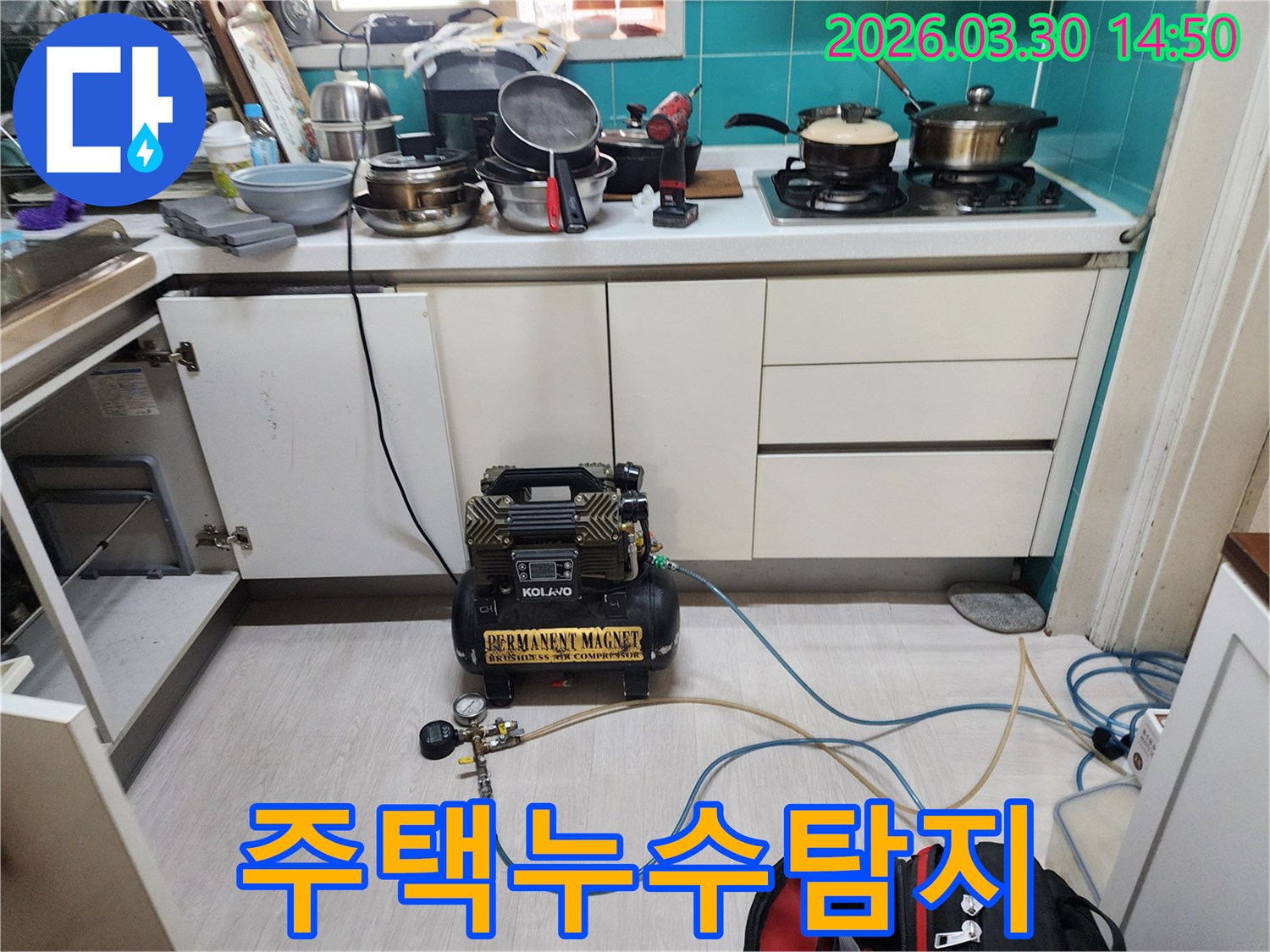 주방 하부장 아래에 위치한 배관에-누수 탐지용 공압 장비가 연결되어-압력 테스트가 진행되고 있습니다.