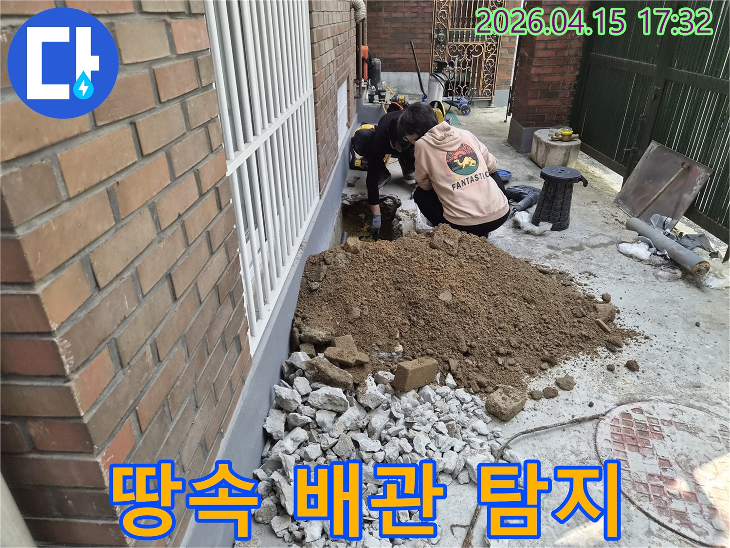 두 명의 작업자가-건물 옆 땅을 파내어-누수된 배관을 찾아내고 있는 현장