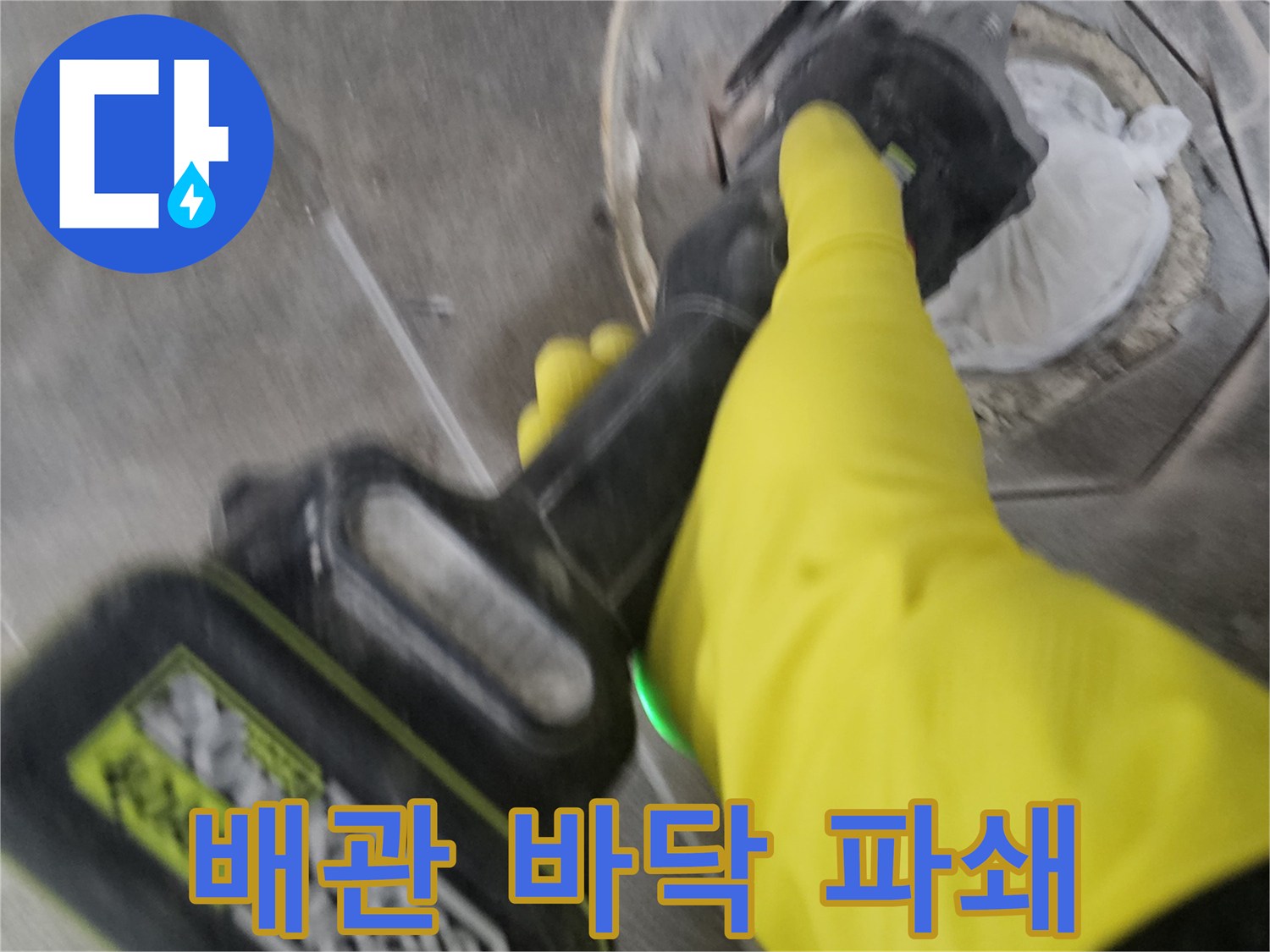 노란색 보호 장갑을 낀-전문가가-전동 절단기로-배관 주변의 바닥 타일을-조심스럽게 파쇄하는 장면입니다.
