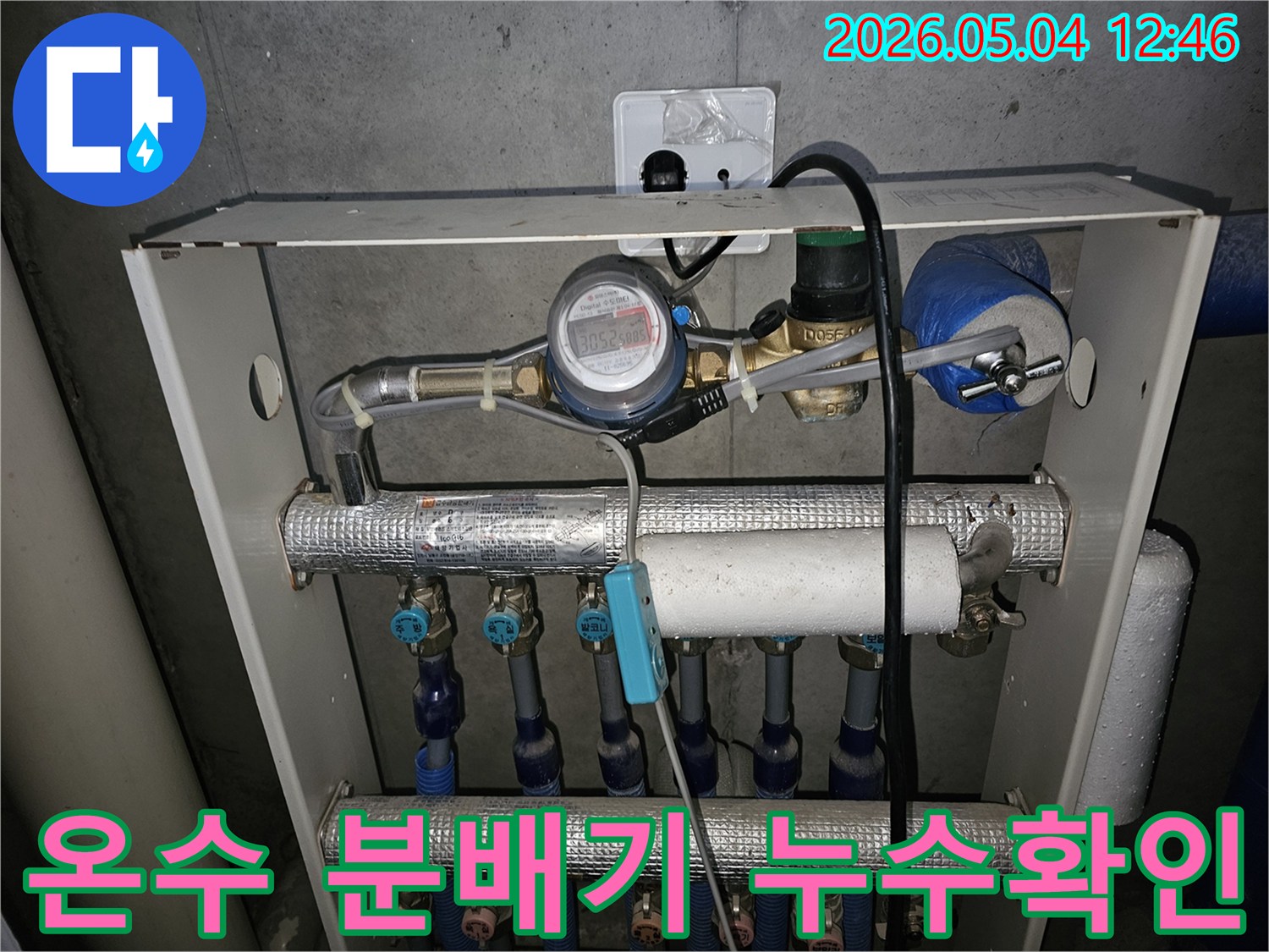 이 이미지는 아파트 온수 분배기 주변에서 누수 여부를 확인하는 장면입니다-배관과 유량계가 선명하게 보입니다.