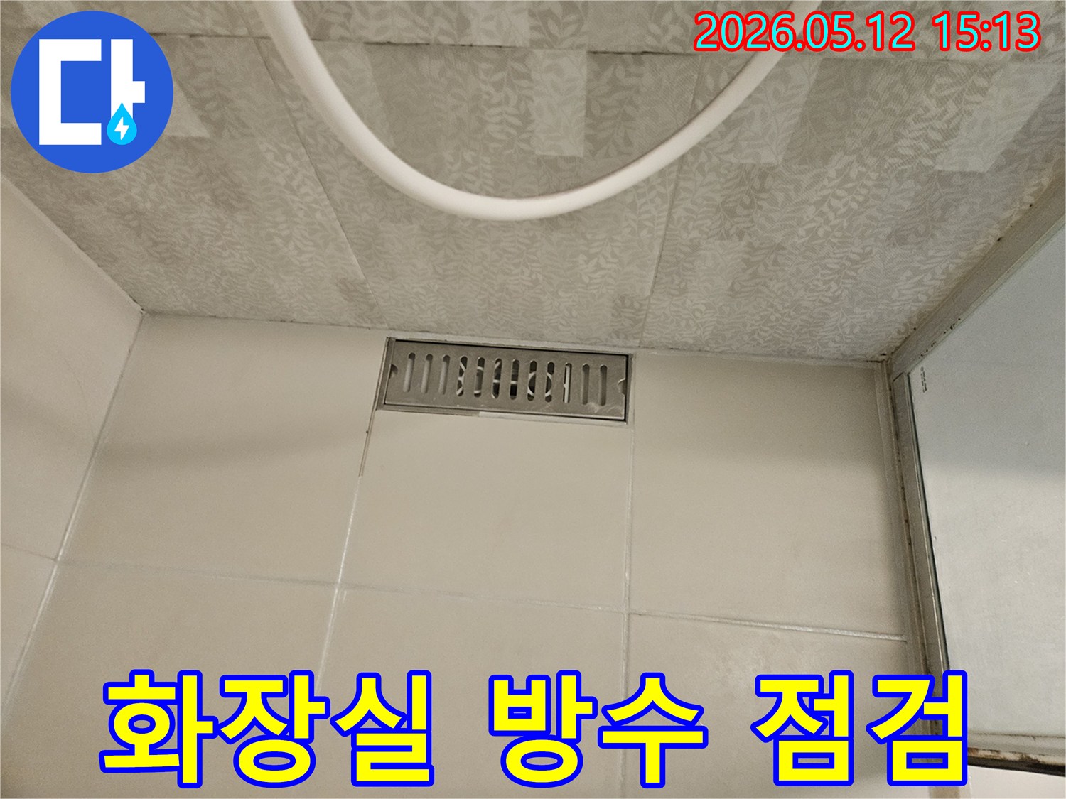 화장실 바닥의 직사각형 스테인리스 배수구와-주변의 흰색 타일-그리고 천장 마감재가 보이는-누수 점검 전 현장 사진입니다.