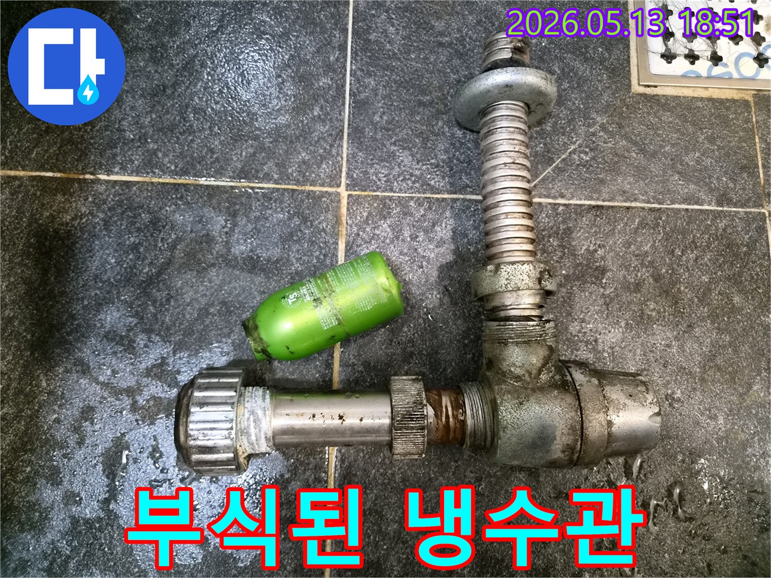 어두운 타일 위에-부식으로 손상된 냉수 배관 부품과-초록색 샴푸통이 놓여있는-누수 수리 현장 모습