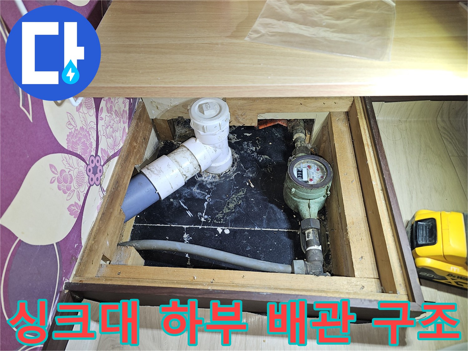 주방 싱크대 아래에 위치한 수도 계량기와 노출된 배관의 모습입니다.