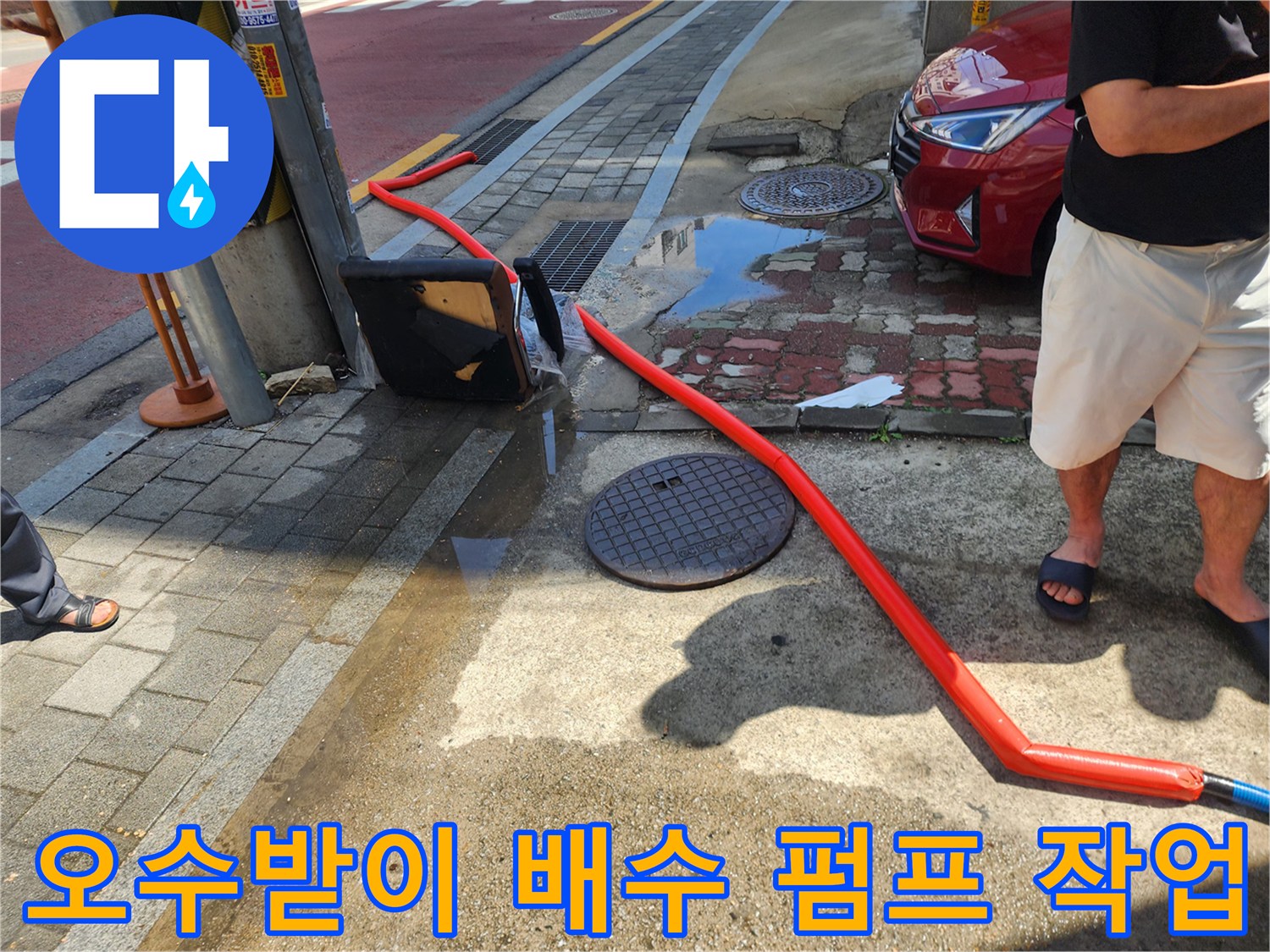 도로변 맨홀 주변에 고인 물을 제거하기 위해 주황색 호스를 연결하여 배수 펌프 작업을 진행하는 모습입니다.