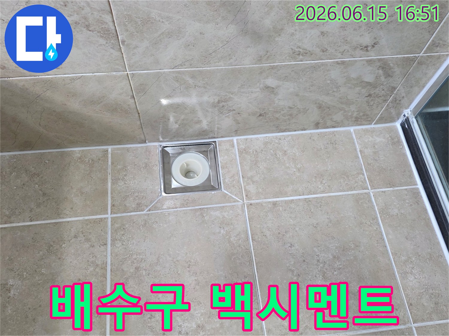 베이지색 타일 바닥에 설치된-사각형 금속 배수구의 모습으로-주변 백시멘트의 마감 상태를 보여줍니다