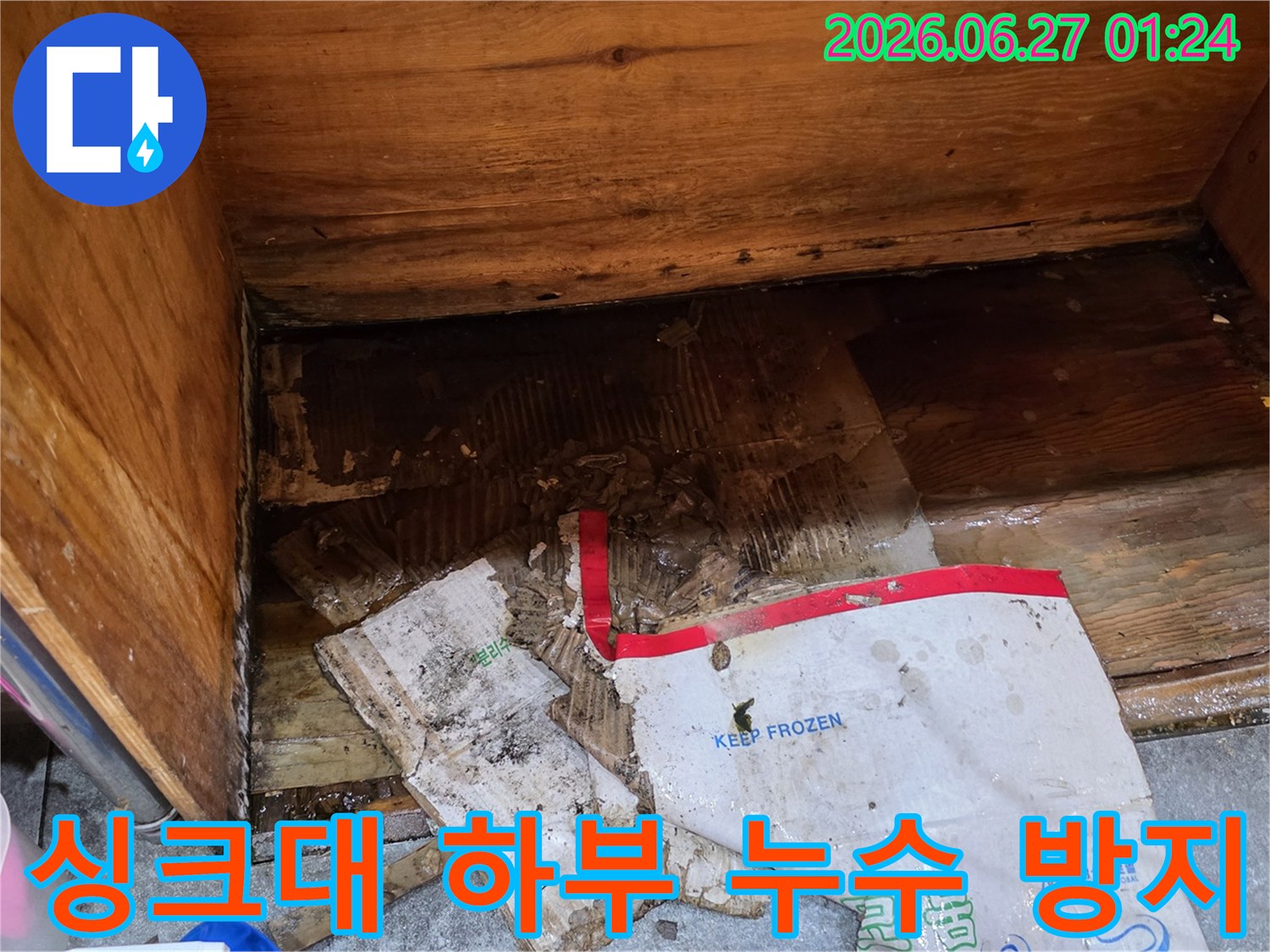 물에 젖어-심하게 손상된-종이 상자와-습기로 인해-변색된 나무가-보이는-주방 싱크대 하부의-누수 공사 현장입니다.