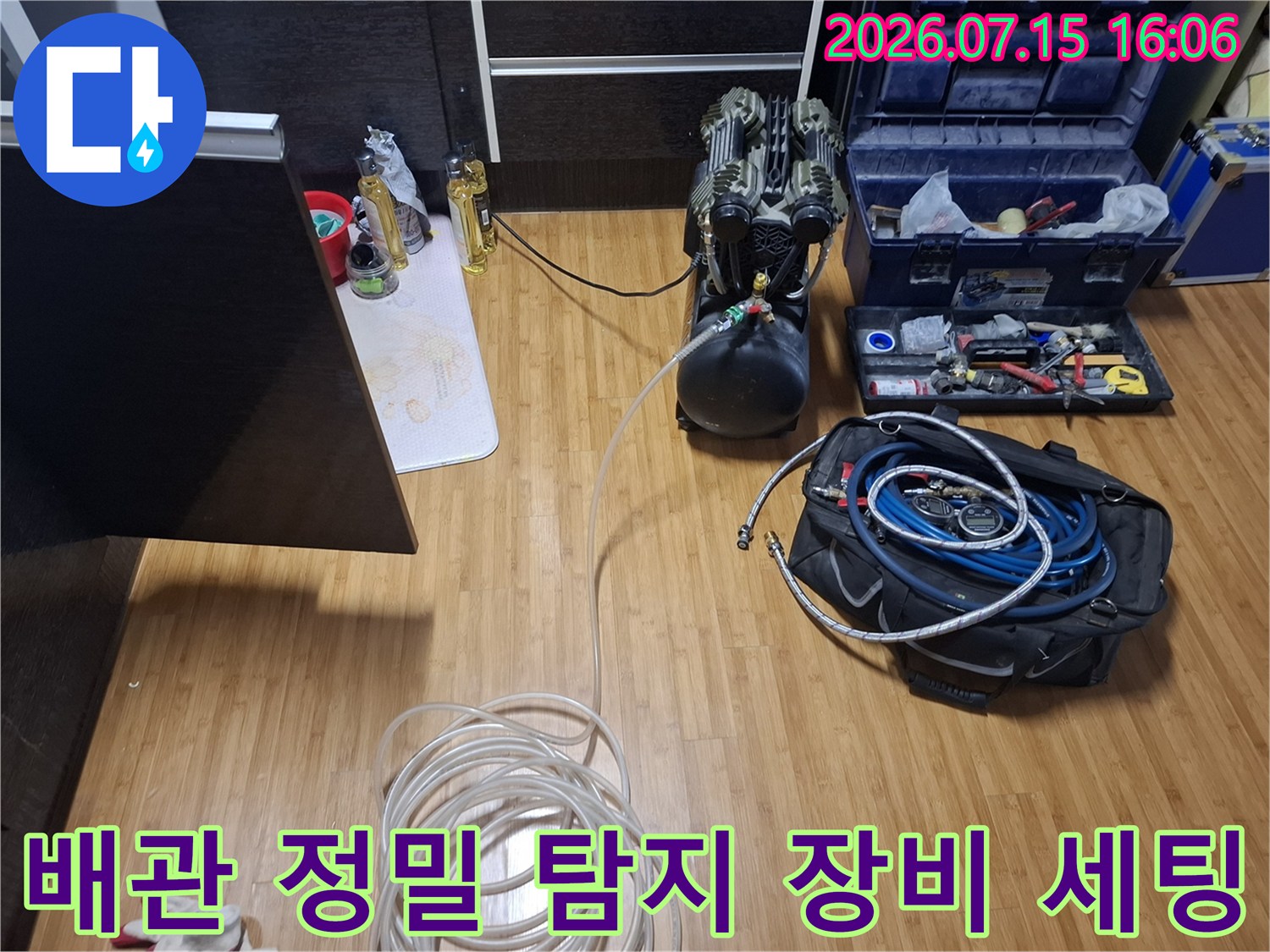 거실 바닥에 배치된 검정색 공압 테스트기-파란색 호스와 공구함-흰색 배관 라인