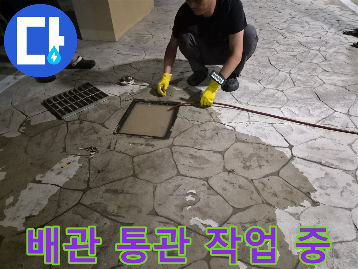 필로티 구조 건물 바닥 맨홀을 통해 붉은색 케이블 장비를 투입하여 배관 막힘을 해결하는 모습