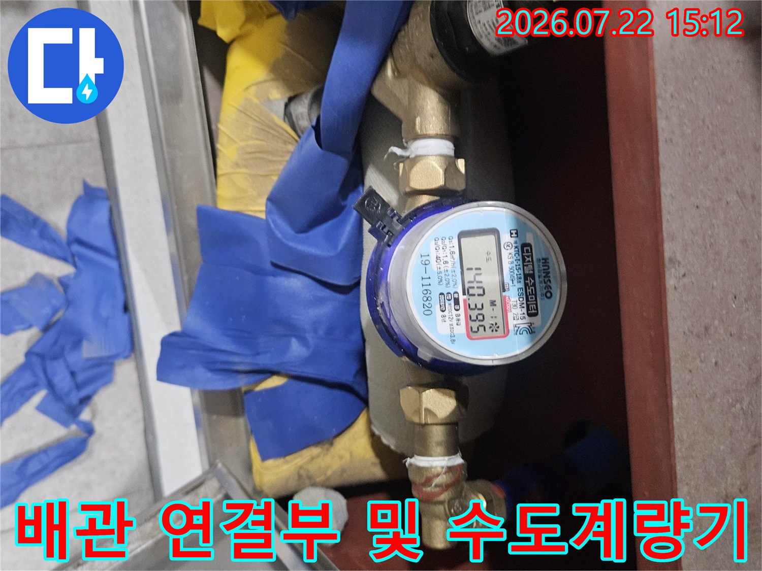 금속 밸브와 파란색 테이핑 처리된 배관 사이에 위치한 수도 계량기의 모습
