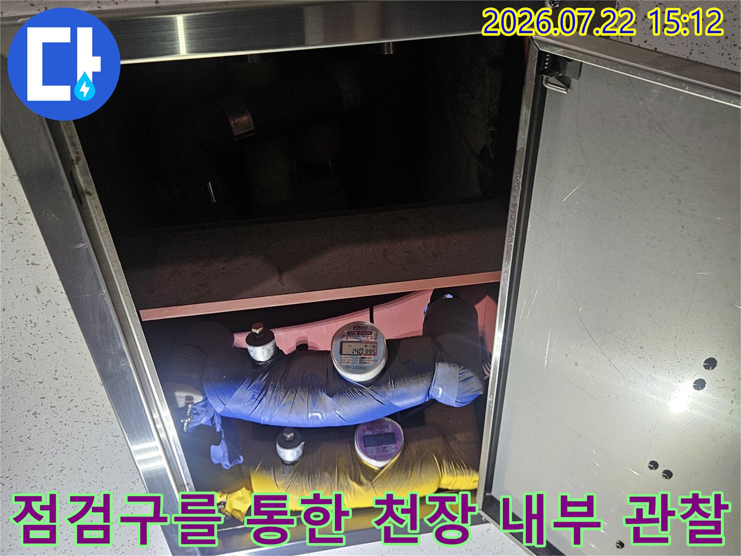 흰색 천장 판넬의 점검구가 열려 있고 그 안으로 보온재가 감싸진 배관들이 보이는 장면