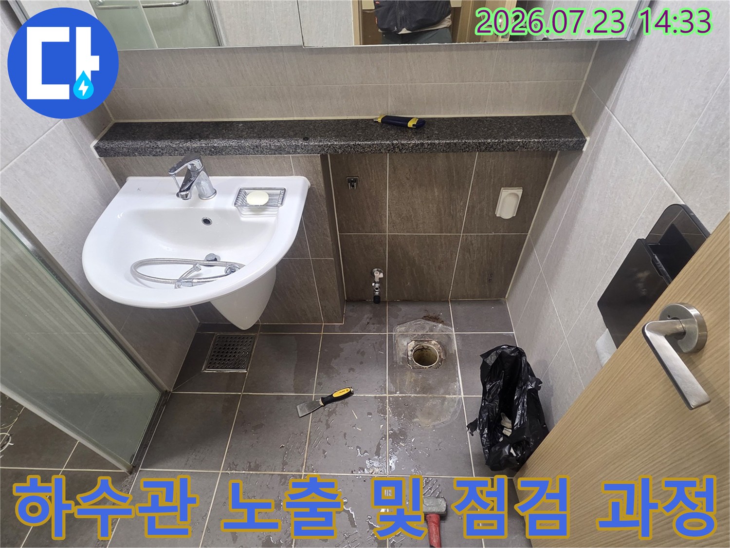 그레이 톤의 욕실 타일 바닥에 세면대가 설치되어 있고 그 아래 배수구 주변이 파헤쳐진 모습