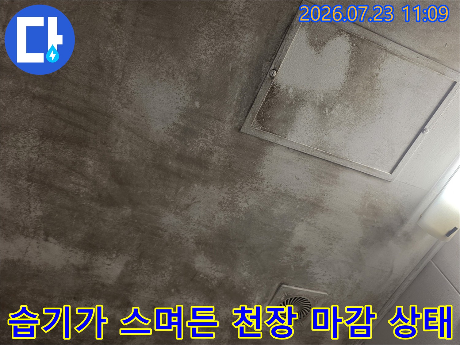 회색빛의 거친 천장 표면에 넓게 퍼진 누수 얼룩과 사각 점검구-환풍기 포함
