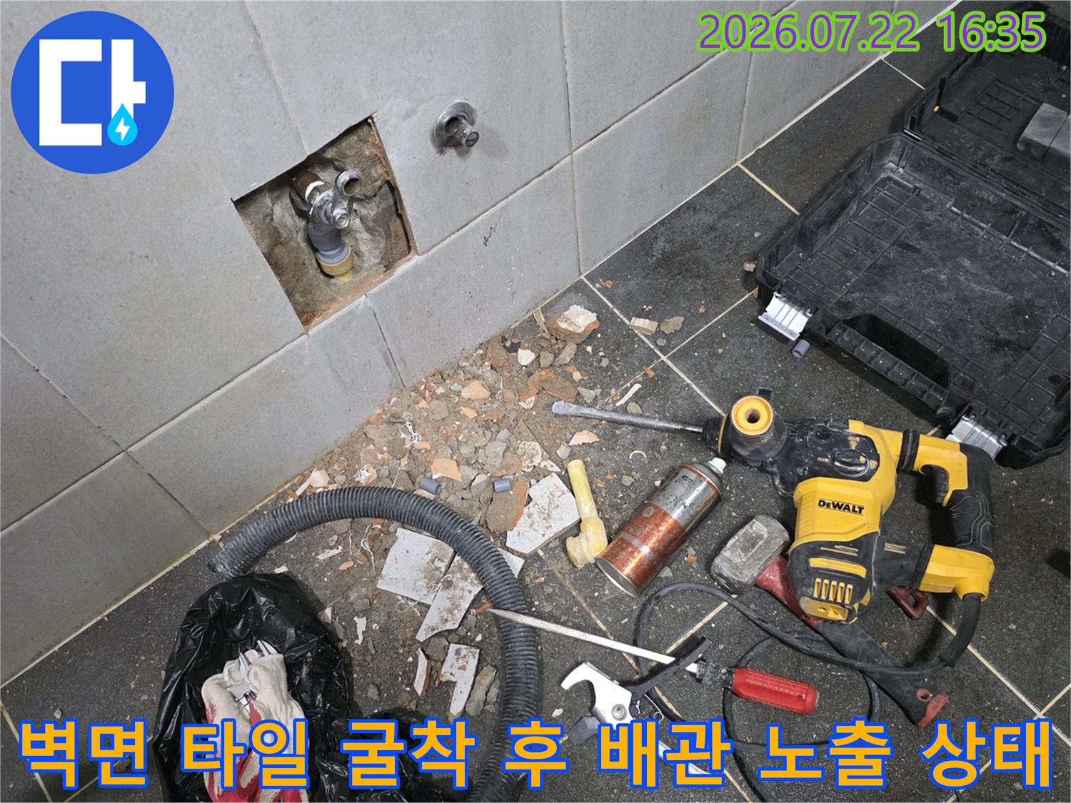 타일 벽면을 굴착하여 내부의 금속 수도 배관과 밸브가 드러난 모습-주변에 전동공구와 장비들이 놓여있음