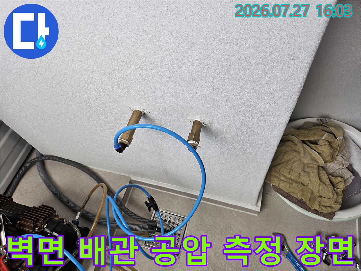 흰색 벽의 배관 연결부에 파란색 호스와 압력 장비가 연결된 상태