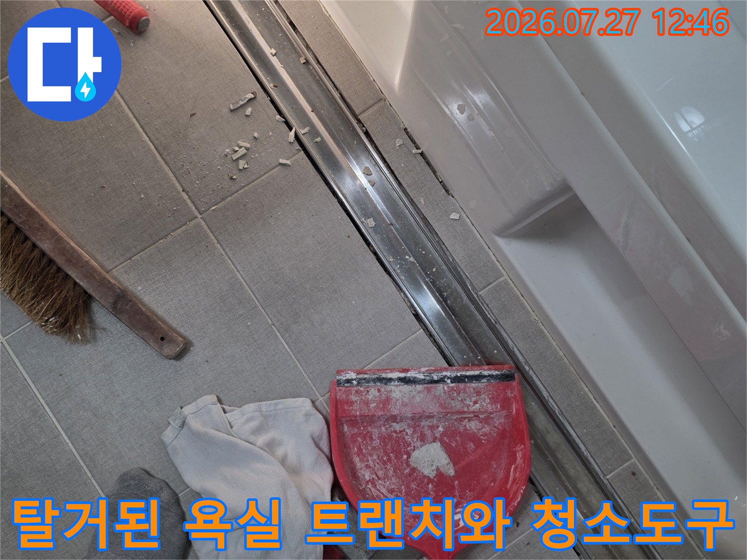 욕실 바닥의 금속제 트랜치가 제거되어 있고 주변에 빨간색 쓰레받기와 빗자루-걸레가 놓여 있는 모습