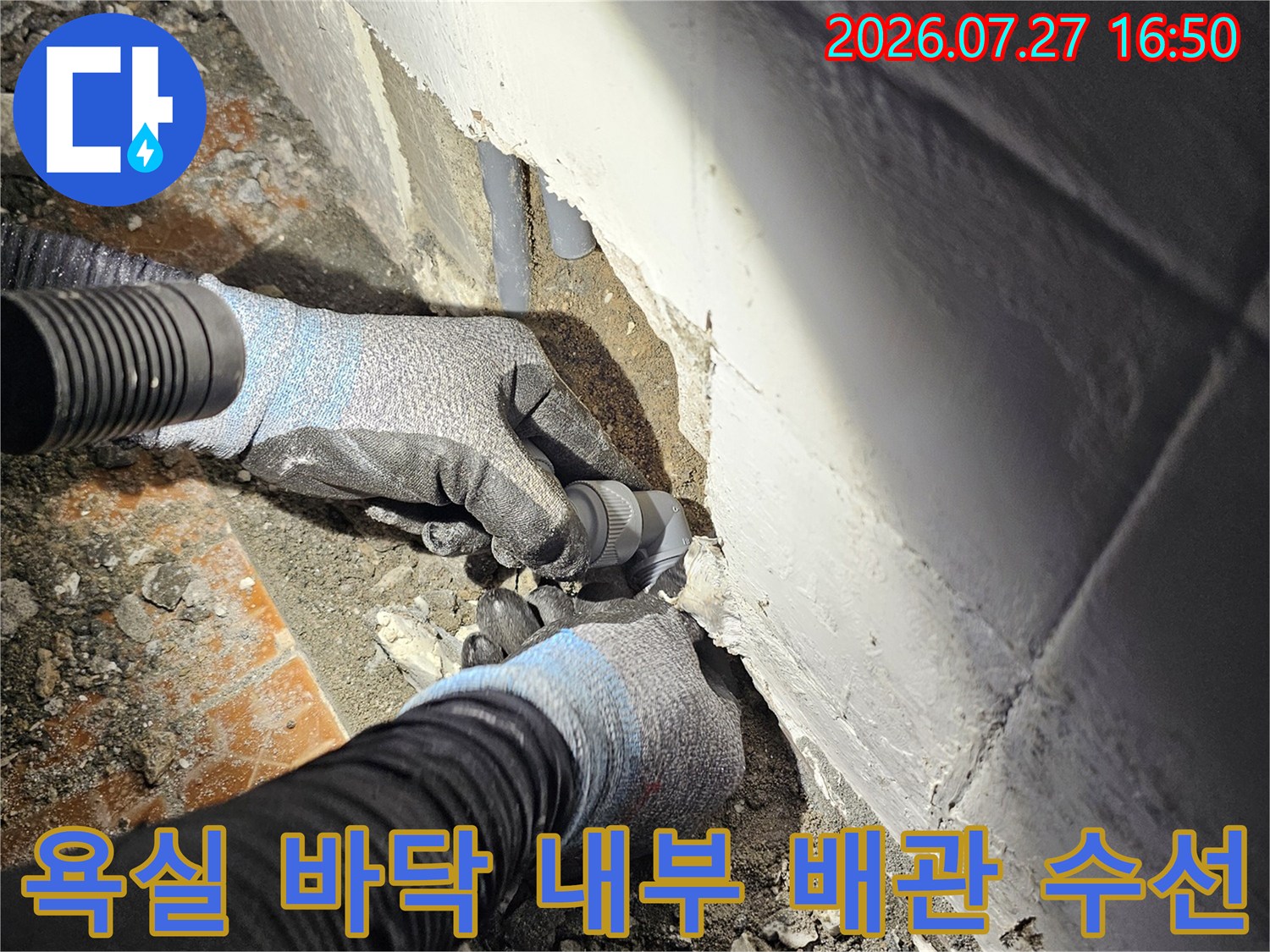 작업용 장갑을 낀 손이 타일이 제거된 바닥 속 회색 배관의 연결부를 조절하고 있는 모습