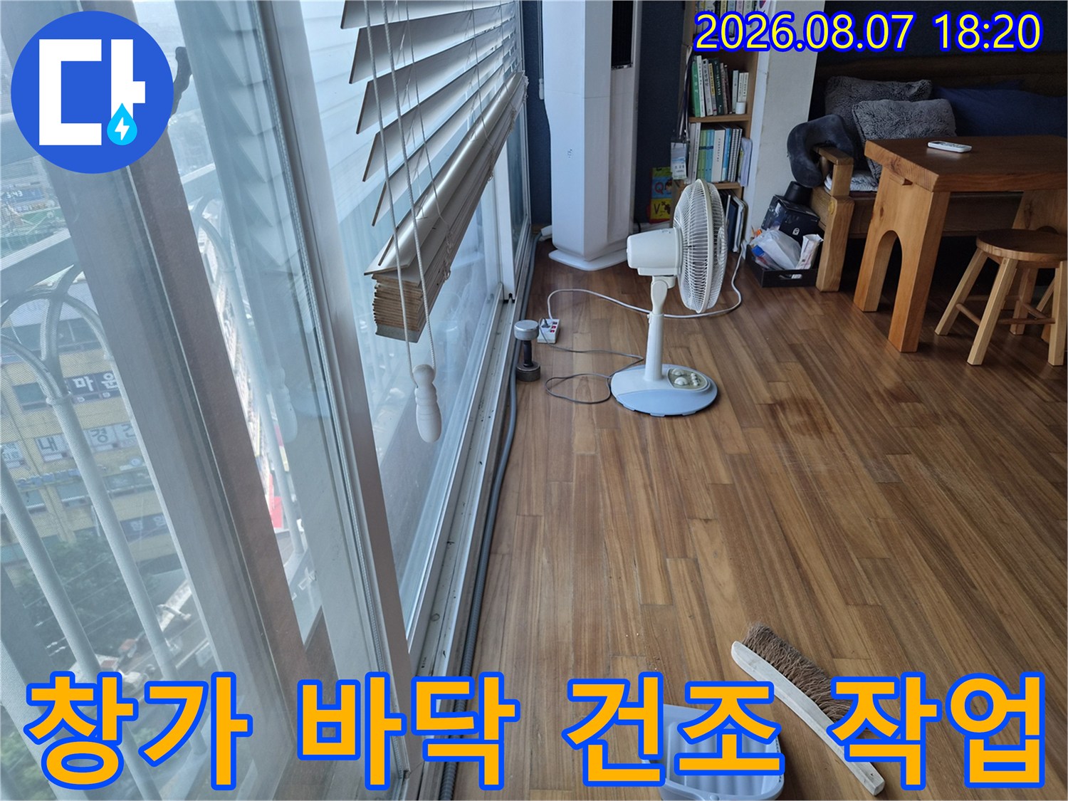 선풍기와 빗자루를 사용하여 누수 흔적을 정리하고 있는 거실 창가 모습입니다.