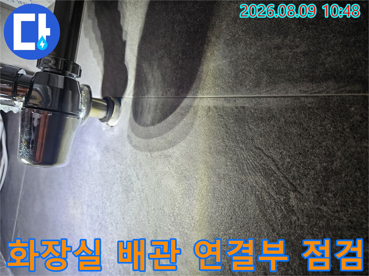 어두운 욕실 타일 벽면에 설치된 은색 배관 부위 근접 촬영