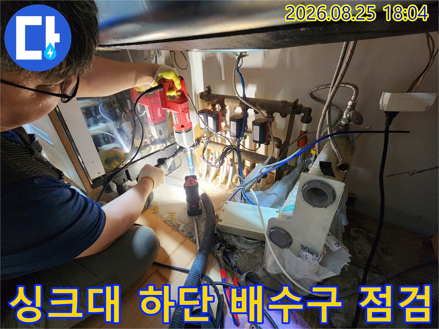 어두운 싱크대 하부장 안쪽의 배관 틈새를 밝은 조명과 장비로 확인하는 작업 장면입니다.