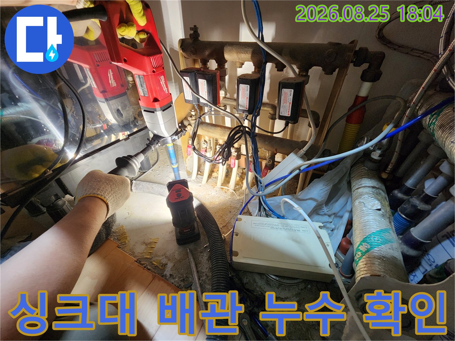 작업자가 드릴을 이용해 싱크대 하부의 복잡한 수도 배관들을 면밀하게 조사하고 있습니다.