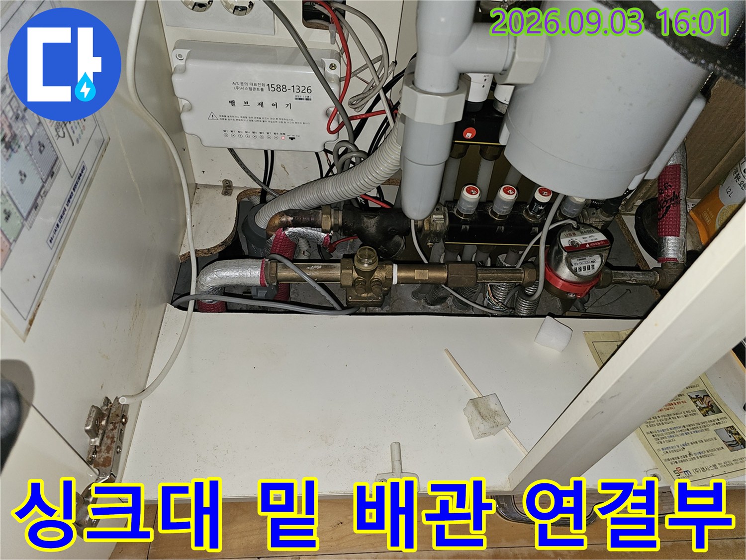 복잡하게 얽힌 보일러 난방 배관과 각종 밸브가 노출된 싱크대 하단 모습입니다.