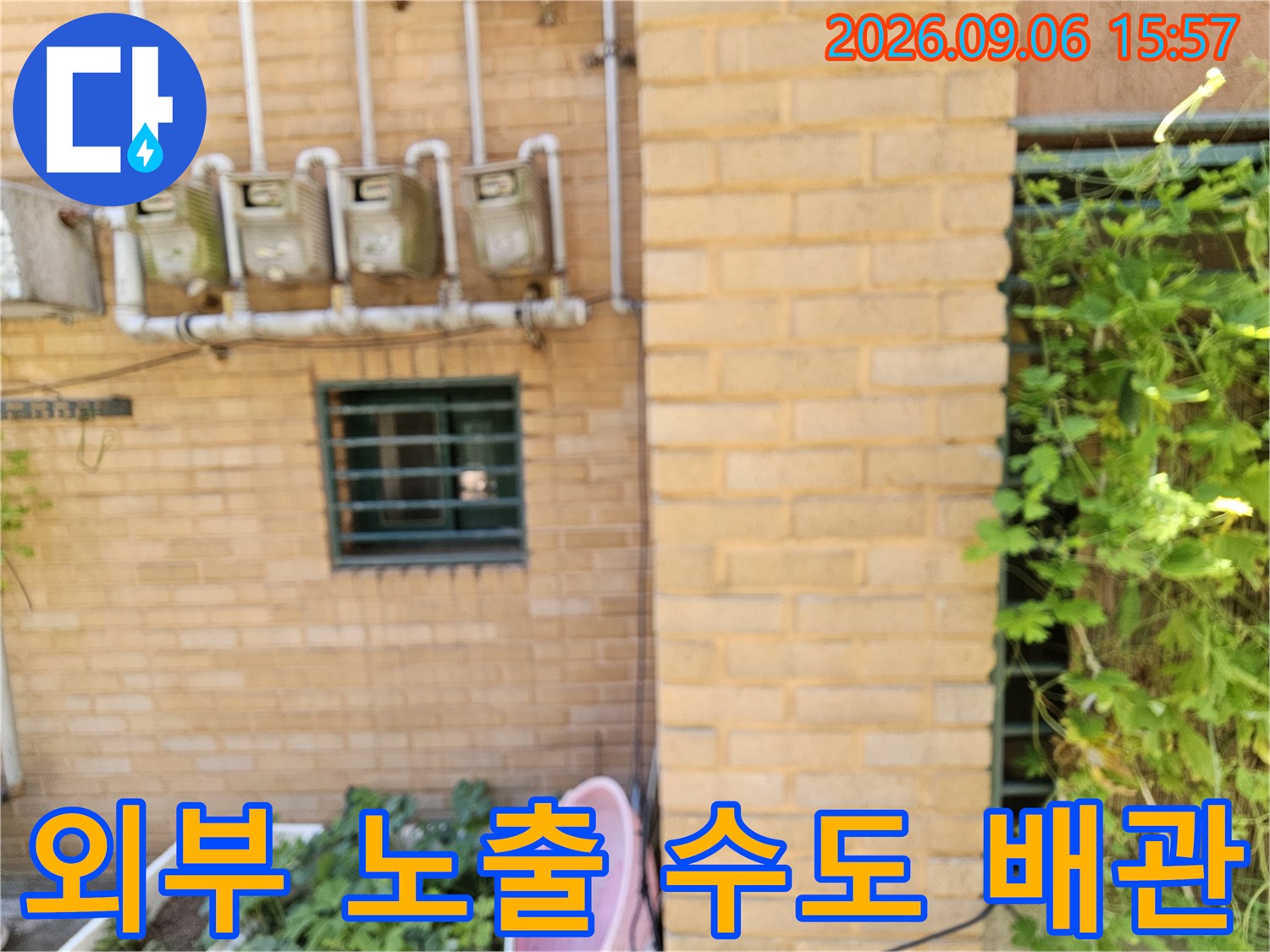 황토색 벽돌 외벽에 설치된 외부 수도 계량기와 배관 연결부입니다.